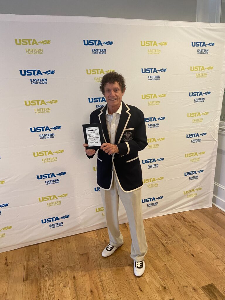 A smiling Marshall Hubsher stands proudly holding his award at the Long Island Tennis Awards dinner, surrounded by fellow tennis enthusiasts. The text reads, "Marshall Hubsher getting award at Long Island Tennis Awards dinner for being #1 on Long Island in Mens 70 and over category."