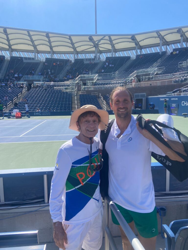 Marshall Hubsher in a colorful tennis outfit and hat, posing with Tennys Sandgren, who is holding a tennis bag, at the US Open with stadium seats and partial view of the court in the background.
